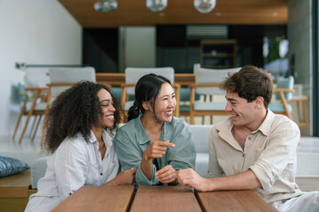 Two young women and a man share laughter at a wooden table, enjoying casual home comfort in light attire, reflecting friendship and diverse cultures.