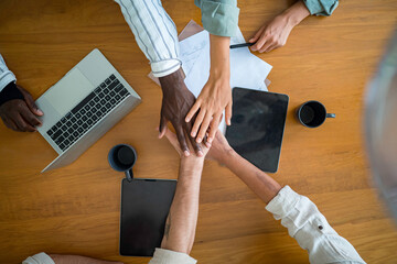 A group of young diverse business executives stack hands in solidarity over a wooden table, surrounded by laptops, tablets, and coffee mugs, symbolizing teamwork in a modern office.