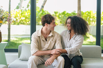 An African American woman and a Caucasian man share a tender moment, sitting closely on a sofa, with joyful expressions, in a bright room.