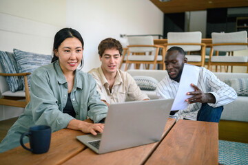 Obraz premium Three young adults of varied ethnicities engage in a collaborative work session in a cozy office lounge area, using a laptop and discussing documents.