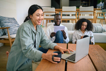Obraz premium Content Asian female adult is focused on her laptop, surrounded by diverse colleagues in a casual, well-lit home office setting, reflecting a modern, collaborative work environment.
