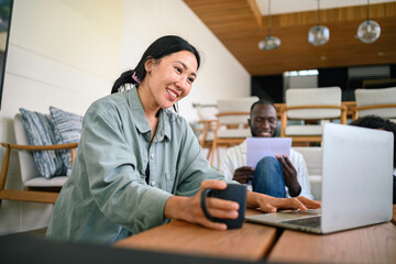 Obraz premium A cheerful Asian female adult works on her laptop, dressed casually in her comfortable home office, sharing a diverse workspace with colleagues.