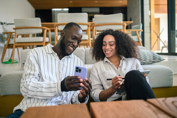 An African American woman and a Black man engage in a work discussion, using a smartphone and digital tablet, seated comfortably in a stylish office lounge.