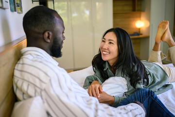 A joyful Black man and Asian woman lounge comfortably in a cozy bedroom, engaging in a heartening conversation, dressed in casual home attire.