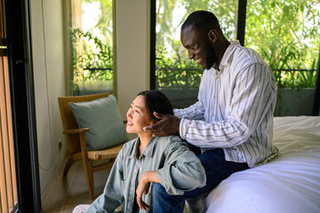 Obraz premium A serene Black man gently massages the shoulders of his Asian partner, both dressed in casual attire, sharing a peaceful moment in a sunlit room at home.