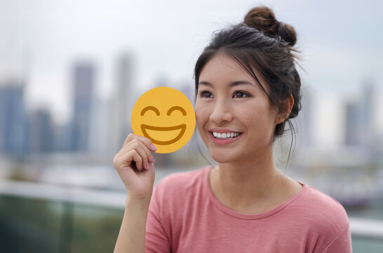 Asian woman cheerfully displays a smiley face cutout, representing positive mental health in a city setting