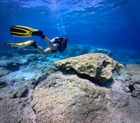 Scuba diver with yellow fins exploring underwater seascape with boulders and stones. Vivid blue...
