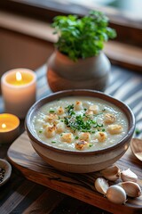 A bowl of creamy garlic soup garnished with croutons and chopped herbs sits on a wooden table. Ambient candlelight and a small plant create a warm and inviting atmosphere