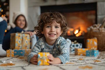 A young child smiles brightly while opening a present, surrounded by colorful gift boxes. A festive fire crackles in the background, creating a cozy holiday atmosphere with family nearby
