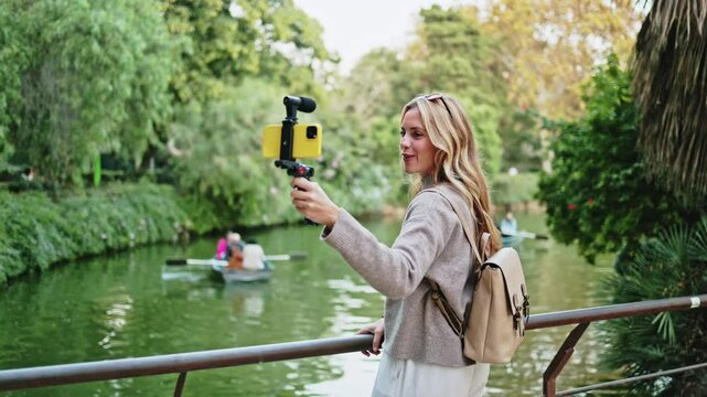 Travel influencer filming a vlog by a lake, capturing moments of people rowing boats in the background.   Travel influencer capturing scenic vlog moments by a serene lake