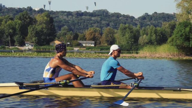 Rowing team training. Side view of 2 young caucasian male rowers, during a rowing practice, athlete sitting in a boat in the river Dnipro, rows through a calm water sunny day, autumn. 4k footage