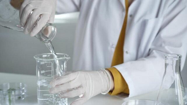 Unknown scientist wearing a lab coat and gloves is carefully pouring a liquid from one beaker to another in laboratory set, close up. Science and medicine concepts