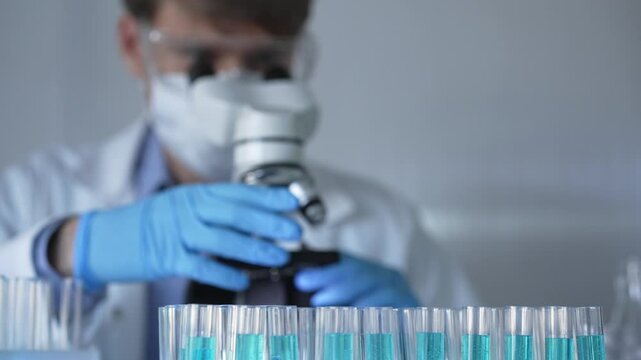 Laboratory tubes are in focus, closeup view. Man scientist researcher with blue protective gloves is using a microscope at the background. Medicine and science concept