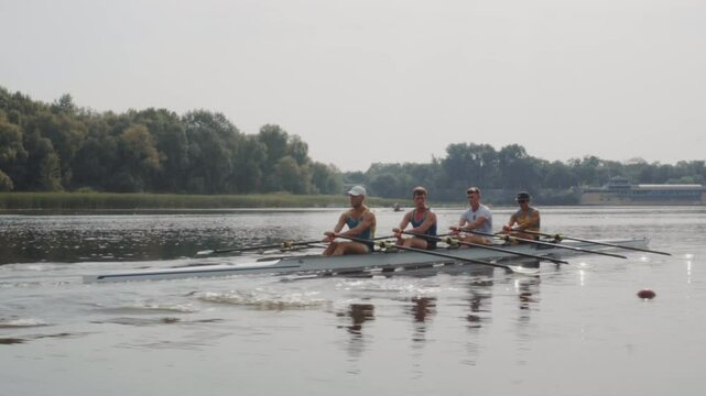 Rowing team training. Side view of 4 young caucasian male rowers, during a rowing practice, athlete sitting in a boat in the river Dnipro, rows through a calm water in autumn. 4k footage. City area in