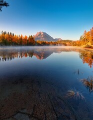 Obraz premium Peaceful Lake at Sunrise, With Mist Rising From the Water and a Lone Mountain Reflecting on the Calm Surface, Surrounded by Autumn Forests in Soft Morning Light