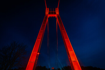 Close-up of a suspension bridge tower lit by red lights against a deep blue night sky, with the...