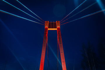 Fototapeta premium Close-up of a suspension bridge tower lit by red lights against a deep blue night sky, with the full moon shining brightly in the background. The cables create dynamic lines, adding visual depth.