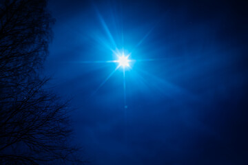 A bright moon casts a striking starburst effect in a deep blue night sky. Bare tree branches frame...