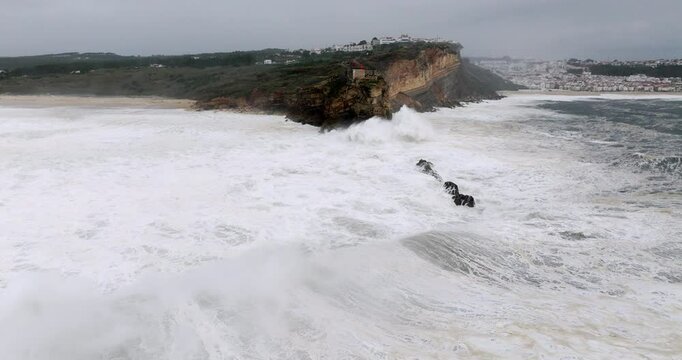 Aerial drone shot of big waves breaking on rocks and cliffs on a day with giant waves in Nazare, Portugal, Europe. Farol da Nazar&eacute; lighthouse visible. Shot in ProRes 422 HQ