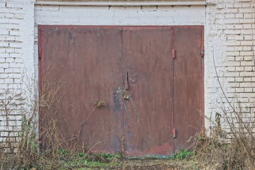 facade of an old garage made of gray brick wall with large closed rusty gate on the street in green...