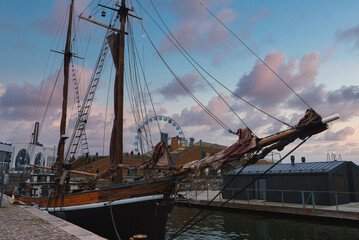 Obraz premium A classic wooden sailing ship is docked at Helsinki harbor, with the SkyWheel in the background. The sunset sky features soft pink and blue hues.