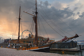 A classic wooden sailing ship is docked at Helsinki harbor, with the SkyWheel in the background. Dramatic clouds and a cobblestone path complete the scene.