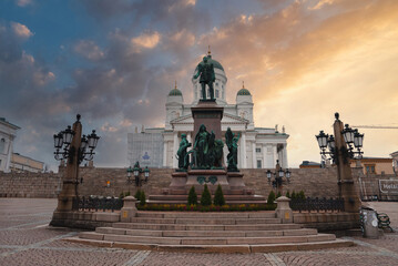 Helsinki Cathedral's neoclassical architecture with white facade and green domes is set against a sunset sky, featuring the Alexander II statue.