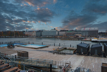 View of Helsinki's waterfront featuring a wooden deck, small structures, and seating areas. The skyline includes a mix of modern and traditional architecture.