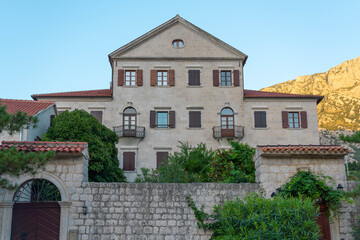 Fototapeta premium A traditional stone villa with wooden shutters and balconies, surrounded by lush greenery and set against a mountainous landscape in the Mediterranean.