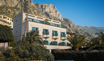 A beautiful Mediterranean hotel with blue balconies, nestled against a mountain backdrop, surrounded by palm trees and lush greenery.