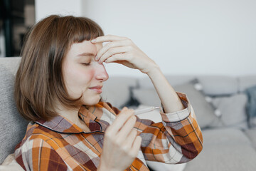 Sick Young Woman In Pajamas Sitting On Sofa Holding Thermometer And Touching Nose Bridge Suffering From Sinusitis At Home