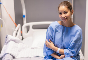 Obraz premium young beautiful woman in private hospital room, dressed in blue robe, sits on bed and rejoices at quick correction of cosmetic defect, waits for doctor's examination before plastic surgery,
