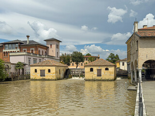 Portogruaor, Venice, Italy - June 26, 2024: Historic yellow stone watermill buildings on Fiume Lemene, greenish river. Waterfall and mansion building on side under blue cloudscape