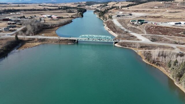 Vehicles travel across a bridge overlooking a river with Canadian Rockies at background aerial revealing shot.
