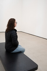 Young Caucasian Woman Sitting on a Bench in a Minimalist White Room, Contemplating