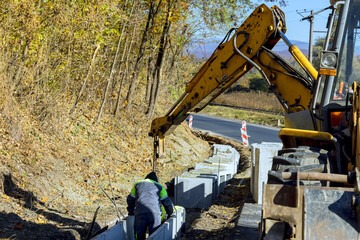 Fototapeta premium Construction worker operates crane to position concrete U blocks beside rural road.