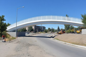 Fototapeta premium A white pedestrian bridge arches over a street under construction with a clear blue sky.