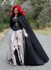 full length portrait of beautiful red haired female model figure, wearing gothic fantasy costume with torn ballgown wedding dress, black corset, leather boots.  walking pose forest scenery background.