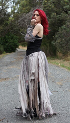 full length portrait of beautiful red haired female model figure, wearing gothic fantasy costume with torn ballgown wedding dress, black corset, leather boots.  walking pose forest scenery background.