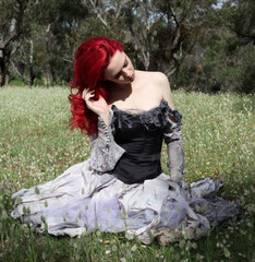 full length portrait of beautiful red haired female model figure, wearing gothic fantasy costume with torn ballgown wedding dress, black corset, leather boots. sitting pose forest scenery background.