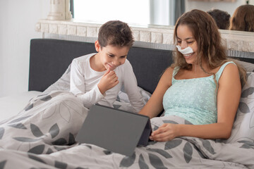 Smiling mother with bondage on her face after rhinoplasty lies in bed with her little son and watches movie on digital tablet,concept of joint leisure