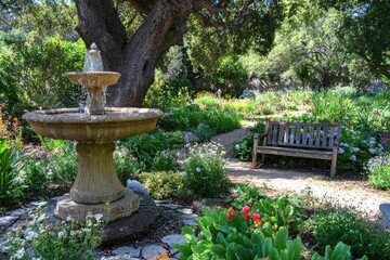 A serene garden featuring a fountain, lush flowers, and a wooden bench for relaxation.