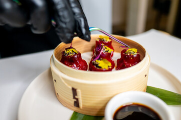 A person wearing a black glove uses golden tongs to pick up a red dumpling from a bamboo steamer....
