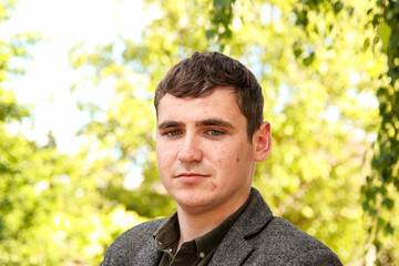 A young man in his 20s standing outside in nature, with visible acne and scars on his face. The...