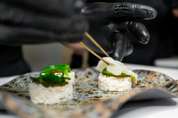 A close-up of a chef’s hands wearing black gloves, meticulously plating sushi. The image...