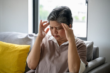 An elderly Asian woman is sitting on a sofa at home, experiencing a headache, dizziness, and a migraine. She looks unwell and uncomfortable. Senior health care daily lifestyle problem concept.