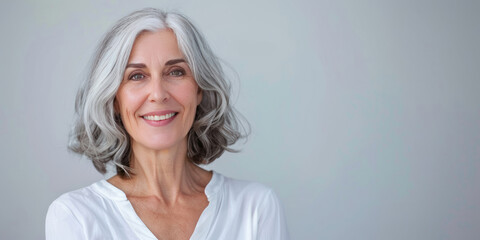 Fototapeta premium An elegant woman in her fifties with silver hair, wearing a white blouse, smiles warmly at the camera against a plain light grey background.