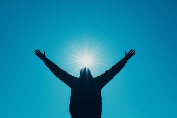 A person standing under a bright blue sky with arms raised towards the sunlight. A concept of freedom, spirituality, and connection with nature.