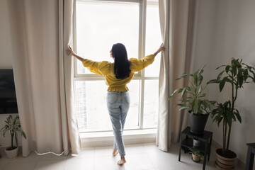 Beautiful black haired young woman standing at large window in stylish cozy apartment, opening...