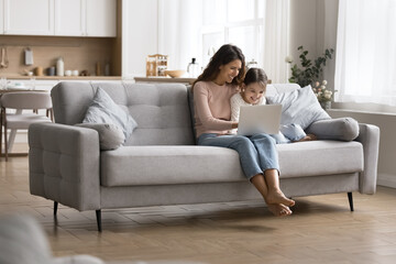 Young mother her little daughter sits comfortably on sofa with laptop in living room, smiling,...